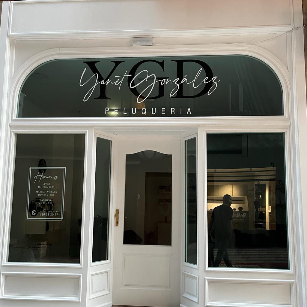 Entrada de la peluquería "Yanet González" con un diseño elegante y moderno, mostrando el nombre del establecimiento en la parte superior y horarios de atención en la ventana.
