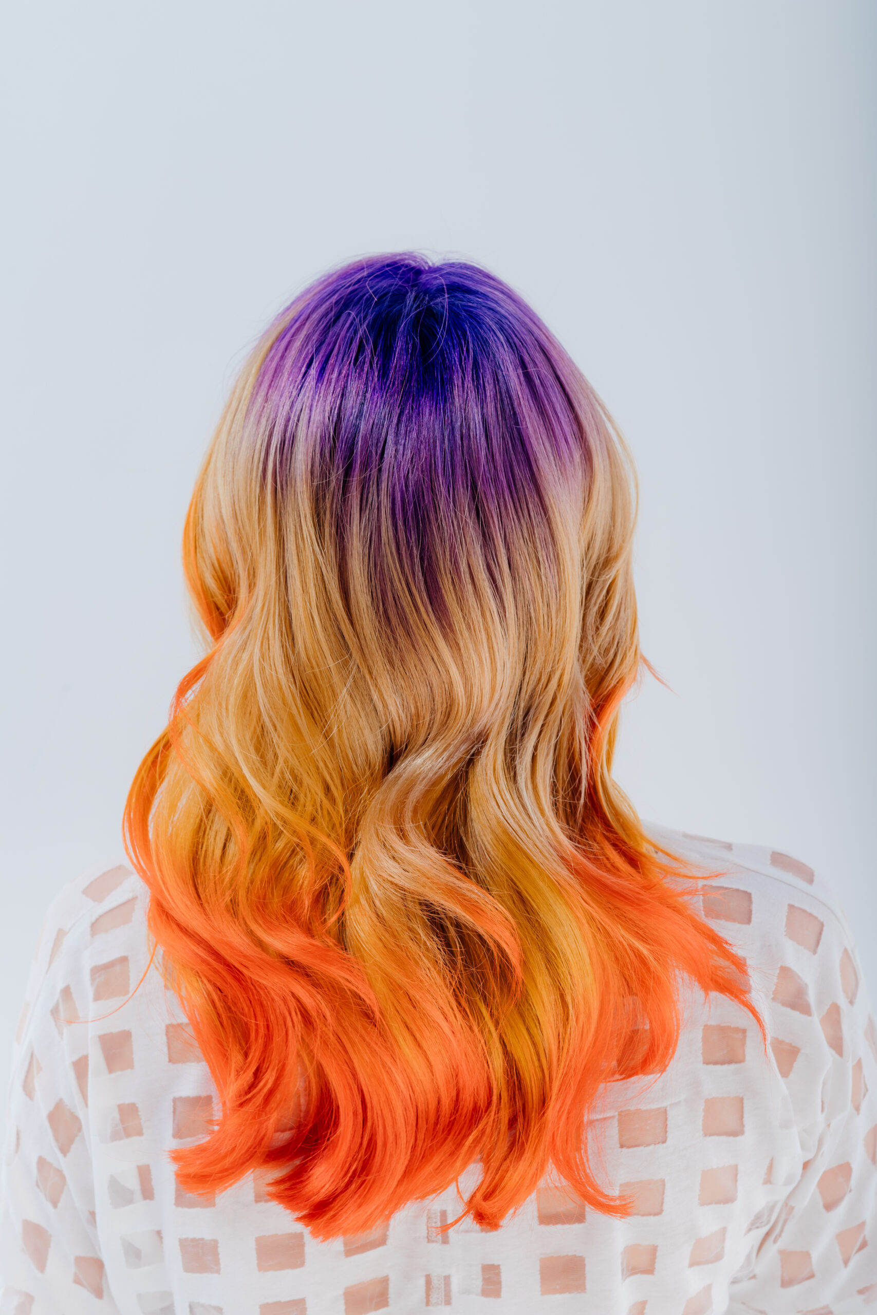 Back view of a person with vibrant, multi-colored hair featuring a gradient from purple at the roots to orange at the tips, showcasing wavy hair texture against a plain white background.